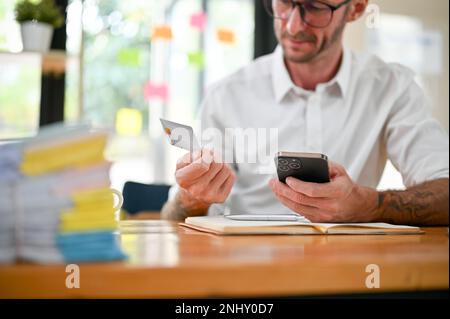 Image rognée d'un homme d'affaires ou d'un comptable du Caucase assis à son bureau à l'aide d'une application bancaire mobile pour payer ses factures en ligne, tenant un credi Banque D'Images