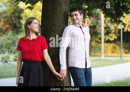 Jeune couple amoureux debout près d'un arbre dans le parc, tenant les mains et regardant l'un l'autre. Banque D'Images