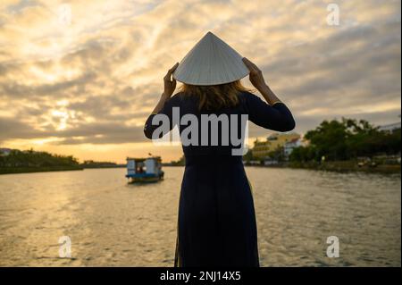 Vue arrière d'une femme méconnaissable avec chapeau conique asiatique et longue robe debout près de la rivière avec eau ondulée et coucher de soleil Banque D'Images