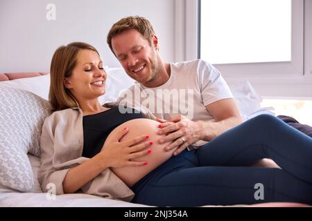 Couple aimant à la maison allongé sur le lit avec l'homme touchant le ventre de la femme enceinte Banque D'Images
