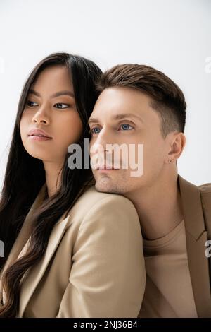 homme élégant se penchait sur l'épaule de la femme asiatique brunette et regardant loin isolé sur gris, image de stock Banque D'Images