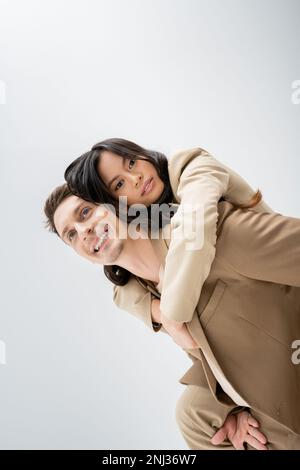 homme souriant dans blazer beige piggysoutenant brune femme asiatique isolée sur gris, image de stock Banque D'Images