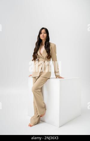 pleine longueur de femme asiatique pieds nus en costume beige debout près du cube blanc sur fond gris, image de stock Banque D'Images