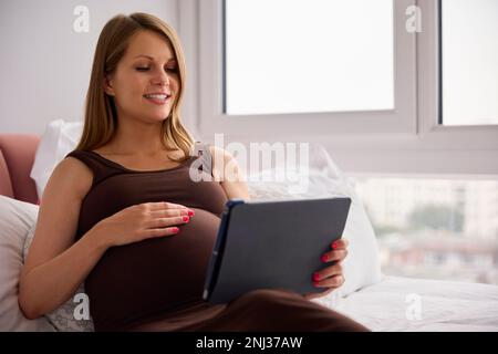 Femme enceinte se reposant sur un lit à la maison regardant la tablette numérique tenir le ventre Banque D'Images