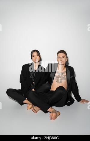 couple interracial pieds nus dans des pantailleurs élégants noirs assis avec des jambes croisées sur fond gris, image de stock Banque D'Images