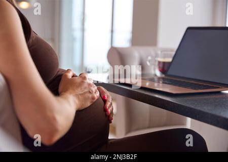Gros plan d'une femme enceinte travaillant à domicile sur un ordinateur portable assis à une table tenant l'estomac Banque D'Images