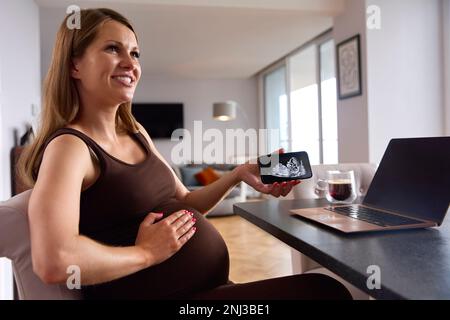 Femme enceinte travaillant à la maison sur un ordinateur portable regardant le téléphone avec l'échographie de bébé Banque D'Images