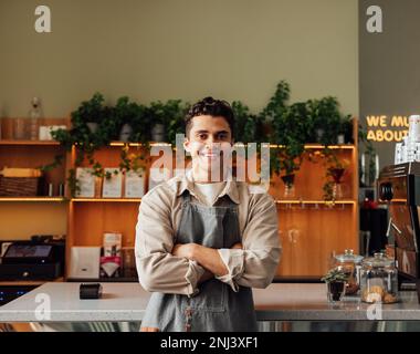 Coffee shop owner in an apron standing with crossed arms and smiling. Young handsome man leaning counter looking at a camera in a coffee shop. Banque D'Images