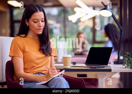 Femme d'affaires assise au bureau dans le bureau pour faire des notes dans le carnet avec une boisson de Takeaway Banque D'Images