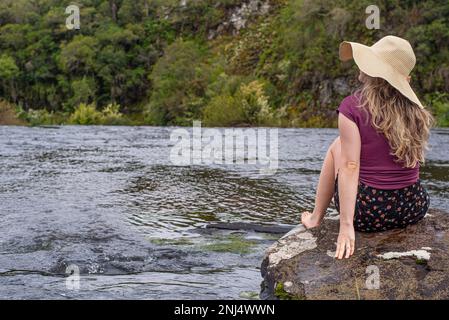 Femme portant un grand chapeau de paille assis sur un rocher sur une rivière Banque D'Images