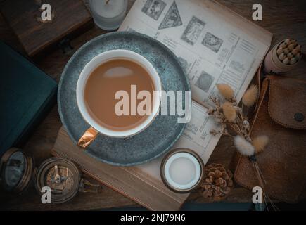 Café chaud dans une tasse en céramique et une cuillère à café en laiton servi avec du lait frais sur une table de travail avec un vieux livre, une boussole en laiton, une bougie parfumée et des fleurs séchées. S Banque D'Images