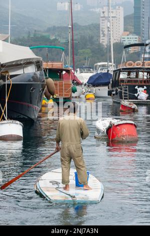 Un homme sur une sorte de stand-up paddle board fait son chemin entre les yachts de luxe dans le port d'Aberdeen, Hong Kong. Banque D'Images