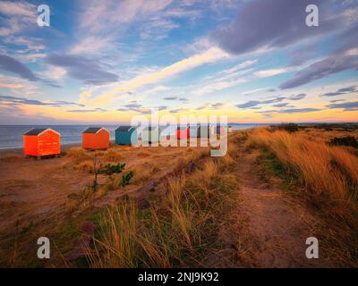 Un sentier sablonneux mène à travers les dunes de Findhorn Beach avec les huttes de plage emblématiques offrant une touche de couleur à mesure que le crépuscule tombe. Banque D'Images