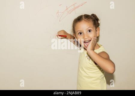 Petite fille brésilienne griffonne, peint et dessinant sur un mur jaune clair Banque D'Images