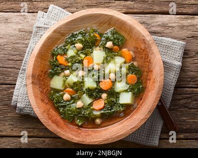 Soupe de légumes frais faits maison à base de chou vert, de pois chiches, de carottes, de céleri, d'oignons et de pommes de terre, servie dans un bol en bois, photographiée au-dessus du bois Banque D'Images