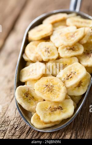 Croustilles de banane croustillantes dans une pelle en métal photographiée sur du bois (mise au point sélective, mise au point au milieu de la puce d'un tiers dans la pelle) Banque D'Images