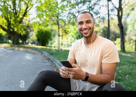 Un jeune sportif afro-américain, athlète, coureur de casque, est assis dans le parc sur le trottoir. Tient le téléphone entre ses mains, écoute de la musique, appelle, se repose. Il regarde la caméra, sourit. Banque D'Images