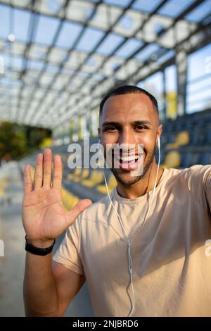 Un jeune athlète afro-américain est debout dans un stade, tient un téléphone et parle sur un appel vidéo par le biais d'un casque. Il regarde la caméra, sourit, dit Bonjour. Photo verticale gros plan. Banque D'Images