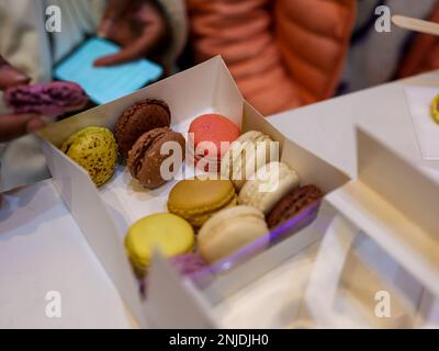 Close-up d'une main plongeant dans une boîte ouverte de macarons pastel colorés disposés en rangées soignées sur une table. Banque D'Images