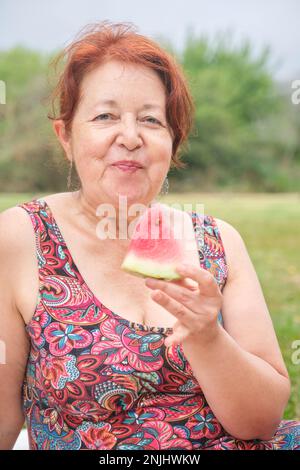 Portrait d'une femme hispanique mûre regardant l'appareil-photo tout en appréciant une tranche de pastèque en plein air, la nourriture rafraîchissante saine d'été. Banque D'Images