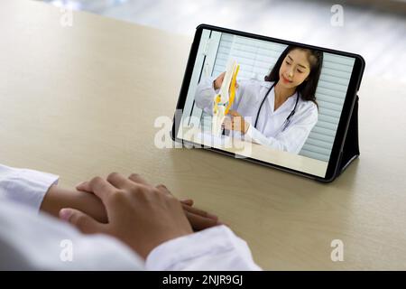 Vue arrière de la femme en robe blanche ont webcam conférence sur ordinateur tablette avec jeune asiatique médecin sur l'os, l'articulation, le tendon et le muscle. Banque D'Images