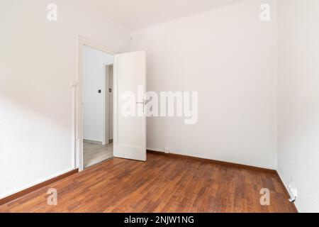 Chambre spacieuse et vide bien éclairée par la lumière du jour. Le sol est en parquet brun foncé. Murs blancs avec une porte donnant sur un couloir. Par l'ouverture Banque D'Images