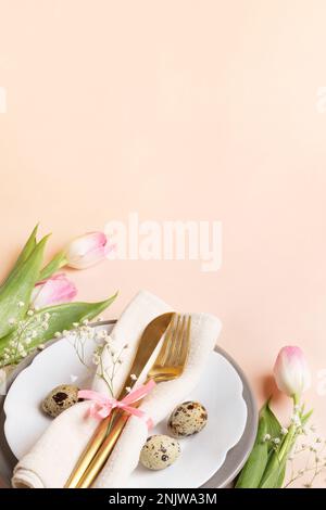 Table de Pâques avec œufs de caille et couverts dorés sur les assiettes, tulipes roses et gitsophila sur fond pêche clair. Composition douce pour les photos Banque D'Images