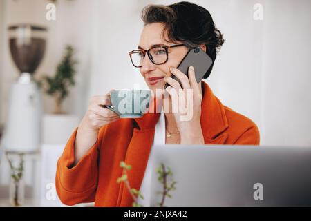femme d'affaires buvant du café tout en prenant un appel téléphonique dans un café. Femme d'affaires mature ayant une conversation téléphonique avec l'un de ses clients pendant WOR Banque D'Images