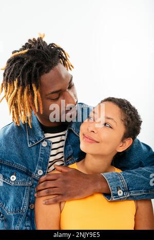 Vue latérale d'un ami afro-américain souriant avec des dreadlocks dans une veste en denim qui embrasse une petite amie avec des cheveux courts qui se regardent l'un l'autre dans un clou Banque D'Images