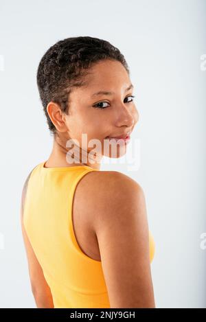 Vue arrière de positive joyeuse jeune afro-américaine avec des cheveux courts en jaune dessus et des jeans debout sur fond blanc regardant l'appareil photo Banque D'Images