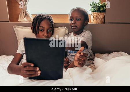 Les enfants noirs sont calmes et se reposent sur un lit sous une couverture et regardent des films ensemble sur une tablette pendant le week-end Banque D'Images