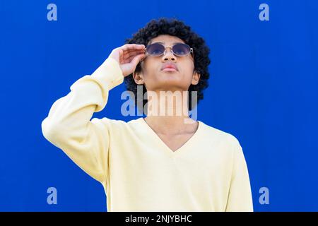 Jeune homme sérieux en sweat-shirt jaune et lunettes de soleil avec des cheveux bouclés regardant vers le haut tout en se tenant contre le mur bleu vif Banque D'Images