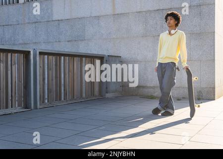 Tout le corps de jeune patineuse ethnique réfléchie de sexe masculin dans une tenue décontractée se penchant sur le skateboard et regardant loin tout en se tenant près de la clôture dans la rue Banque D'Images