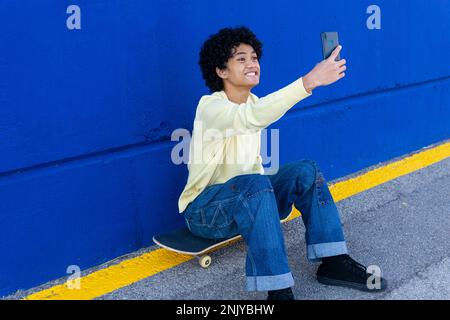 Jeune patineuse asiatique positive en vêtements décontractés souriant et prenant le selfie sur un smartphone tout en étant assis sur un skateboard contre un mur bleu Banque D'Images