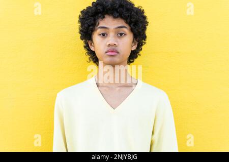 Jeune homme asiatique confiant avec une coiffure afro en sweat-shirt jaune regardant l'appareil photo contre un mur blanc avec ombre au soleil Banque D'Images