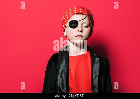 Adorable garçon sans émotion portant un costume de pirate avec les yeux fermés sur fond rouge vif Banque D'Images