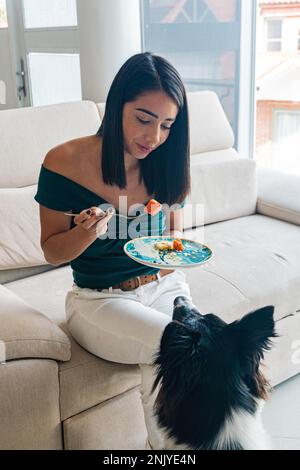 Grand angle de jeune femme hispanique affamée avec des cheveux foncés dans une tenue décontractée assis sur un canapé et manger de la nourriture délicieuse près de curieux adorable Border Collie Banque D'Images