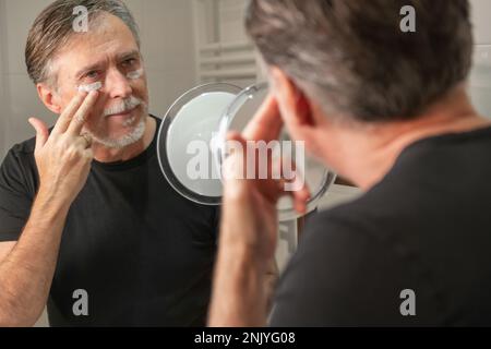 Contenu gris cheveux mâles appliquant de la crème hydratante sous les yeux pendant la routine de soin de peau le matin à la maison Banque D'Images