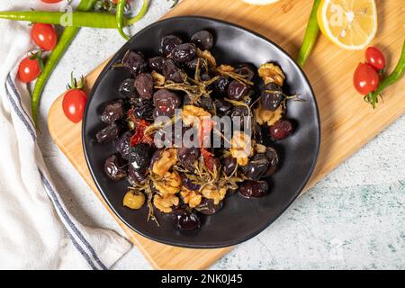Olives noires mélangées. Olives spéciales mélangées préparées avec du poivron rouge séché, des tomates séchées, du thym, des noix et du romarin sur une assiette de service en bois. À Banque D'Images