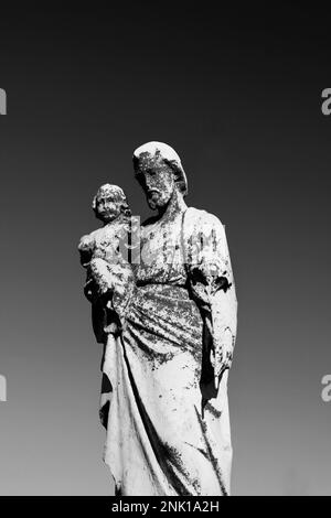 Une statue en pierre naturelle usée et abîmée de Saint Joseph portant le bébé Jésus entre ses mains dans un monochrome noir et blanc. Banque D'Images