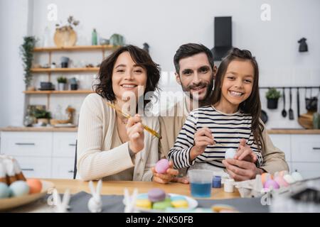 Famille souriante regardant l'appareil photo tout en colorant les œufs Easer dans la cuisine Banque D'Images