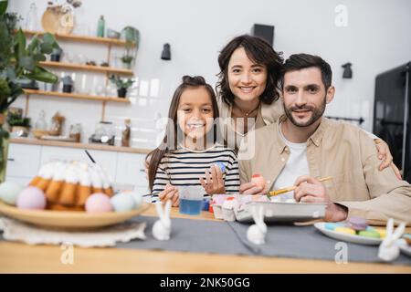 Famille souriante regardant l'appareil photo tout en colorant les œufs Easer dans la cuisine Banque D'Images