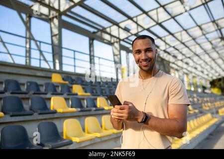 Un jeune athlète afro-américain, entraîneur, coureur portant des écouteurs se tient dans le stade entre les gradins, utilise le téléphone portable. Il regarde la caméra, sourit. Banque D'Images