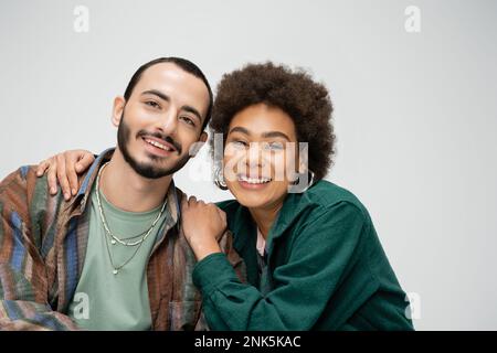 jolie femme afro-américaine souriant à la caméra et embrassant les épaules de l'homme barbu isolé sur gris, image de stock Banque D'Images