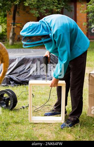 le gardien de faisceau en combinaison de protection recueille les ruches des ébauches en plastique. Récupérer le boîtier de la ruche pour installer les cadres. l'homme tient le foret avec un tournevis et travaille en dehors Banque D'Images