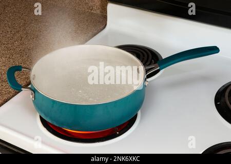 Tir horizontal regardant vers le bas sur une casserole bleue sur un dessus de poêle blanc sur un brûleur chaud rouge contenant de l'eau bouillante. Banque D'Images