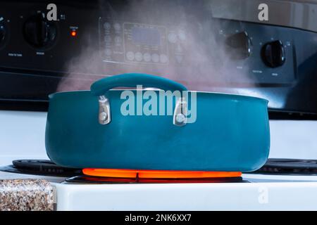 Plan horizontal d'une casserole bleue sur un dessus de cuisinière sur un brûleur chaud rouge avec la vapeur qui s'élève de lui. Banque D'Images