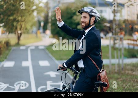 Beau homme élégant en costume bleu marine et casque levant la main pour accueillir quelqu'un sur le chemin du travail. Homme d'affaires caucasien heureux avec pied de vélo noir Banque D'Images