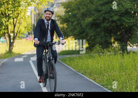 Beau jeune cadre en costume élégant sur la piste cyclable en journée ensoleillée. Vue de face d'un homme d'affaires très occupé portant un casque de protection qui se met au travail Banque D'Images