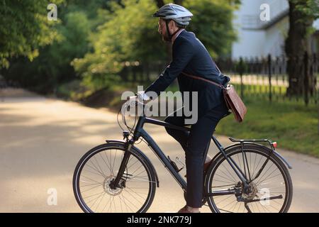 Employé masculin concentré dans un casque de protection sur vélo dans le parc de la ville. Vue latérale du Manager barbu avec porte-documents en cuir, se rendre au travail Banque D'Images